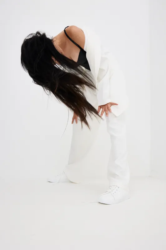 woman bending forward with long hair in white suit and black top, minimalist bright background, dynamic pose expressing movement and energy, modern fashion style