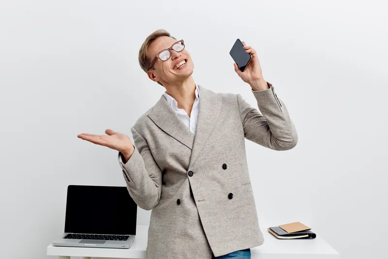 happy caucasian man talking on smartphone wearing glasses gray blazer standing near laptop digital nomad remote work concept