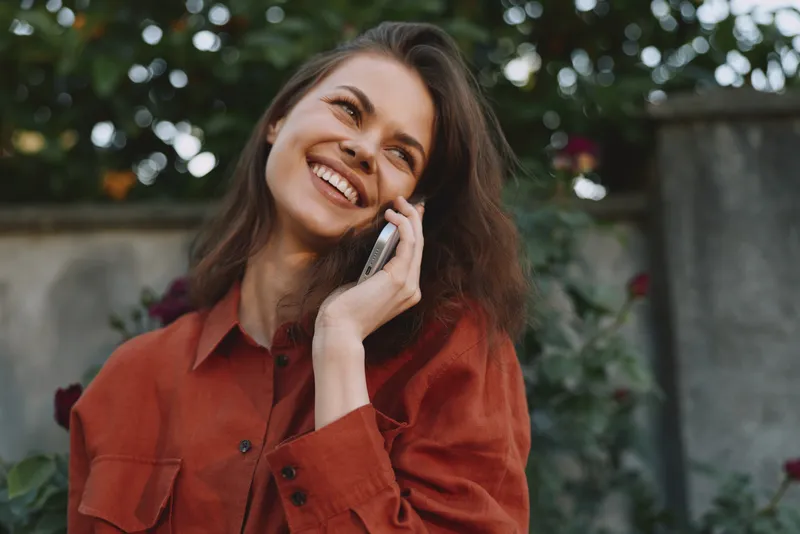 Cheerful woman outdoors, smiling while talking on a phone, relaxed posture, sunlit moment, soft breeze, vibrant greenery background, casual lifestyle portrait
