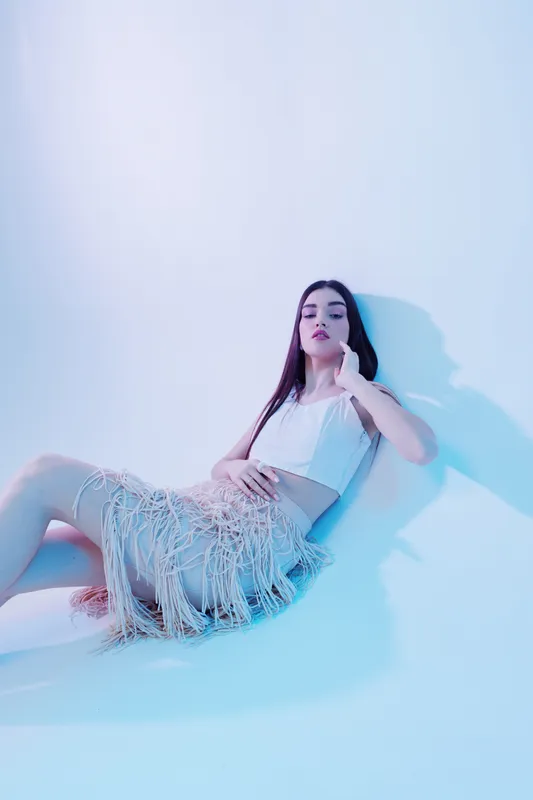 Young woman in a stylish white top and fringed skirt poses sitting on the floor against a light background. Concept of beauty, fashion, and modern style with soft lighting.