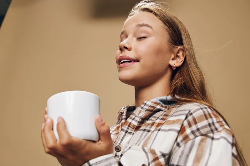 Girl holding white ceramic mug with eyes closed enjoying hot beverage, wearing plaid shirt, relaxing indoors with peaceful expression and cozy atmosphere.