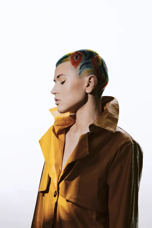 Woman with colorful short hair and artistic hairstyle wearing a shiny brown coat, posing in profile with a calm expression against a white background.