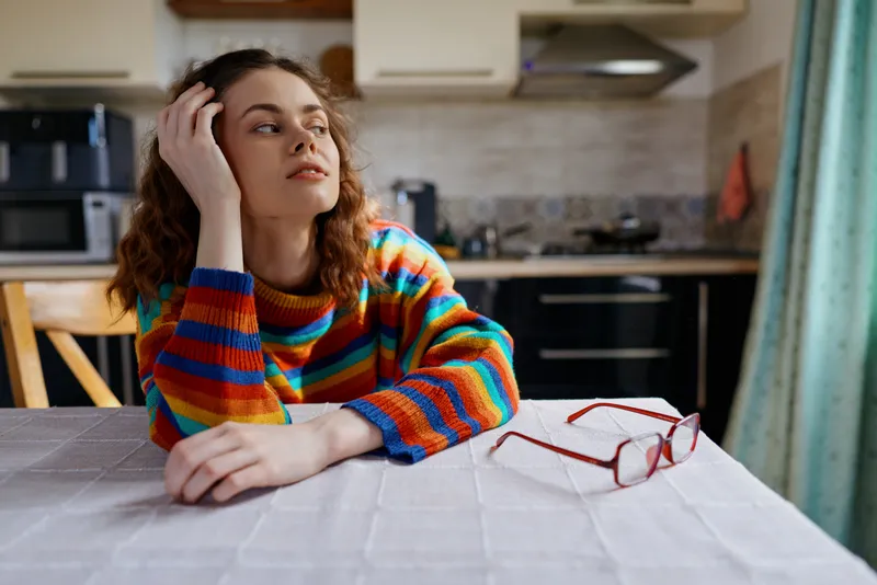 A thoughtful woman sits at a kitchen table, colorful striped sweater, glasses on the table, gazing aside. A candid home life scene conveying reflection, everyday calm, and comfort.