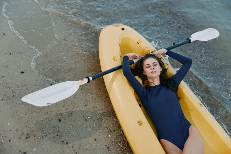 A woman lies in a yellow kayak on a sandy shore, wearing a dark swimsuit, holding a paddle across her chest as gentle waves lap nearby.