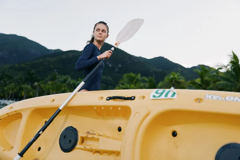 A focused female paddler stands in a yellow kayak, gripping a paddle as she glides on calm water with rugged mountains in the background.