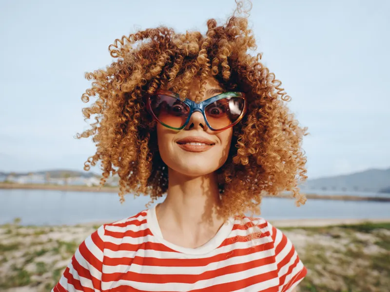Bright portrait of a joyful woman with curly hair, oversized sunglasses, and a red striped tee, outdoors near the coast, with a relaxed smile under a blue sky.