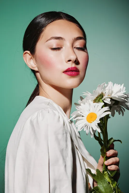 Young woman with glossy hair holds white daisies, closed eyes, wearing bright red makeup and soft white blouse on green background. Creative beauty corner fashion concept.