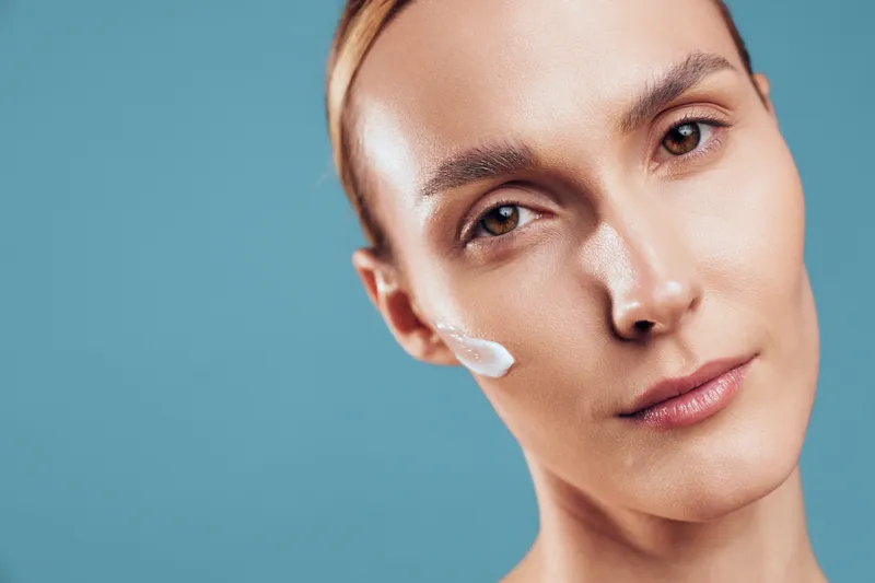 Close up portrait of a woman applying skincare cream, white gel on cheek, calm expression, soft lighting, beauty routine on clean blue background, concept for skin care and confidence.