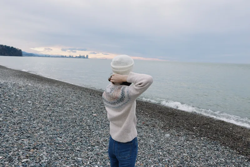 Child standing on a pebbled beach facing the water, wearing a cozy sweater and knit hat, exploring nature and open sky above.