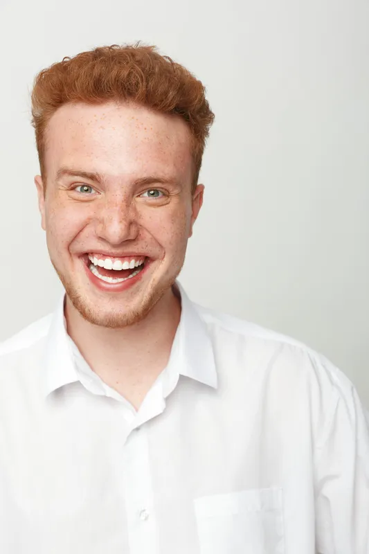 Happy young man with red hair and freckles smiles broadly wearing white shirt. Studio portrait on white background. Concept of joy, positive emotions, youth and casual style.