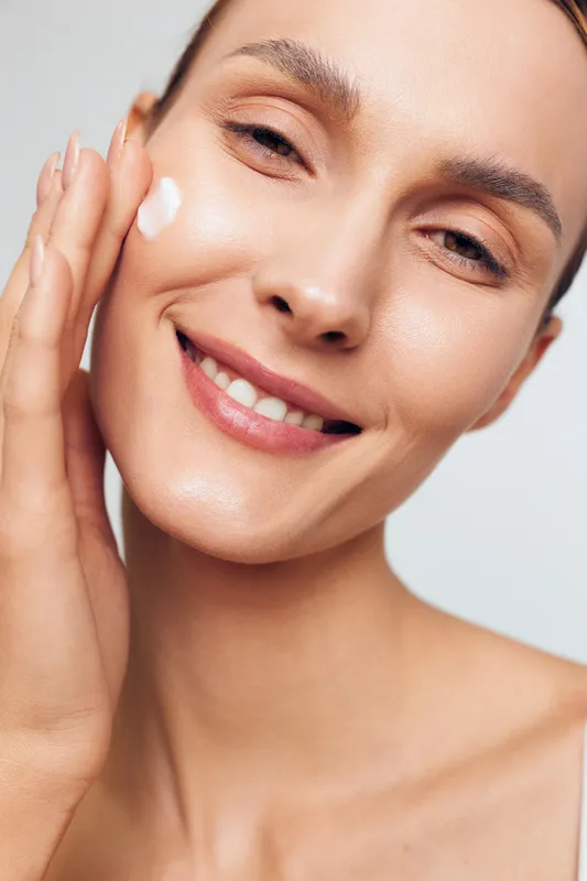 Smiling woman applying cream on face close up, natural skin care routine, fresh complexion, soft lighting, healthy glow, positive mood, beauty treatment concept