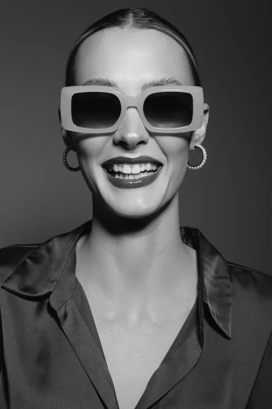 Smiling white woman with fair skin wears large sunglasses and hoop earrings. Studio portrait in black and white with stylish satin blouse and joyful expression.