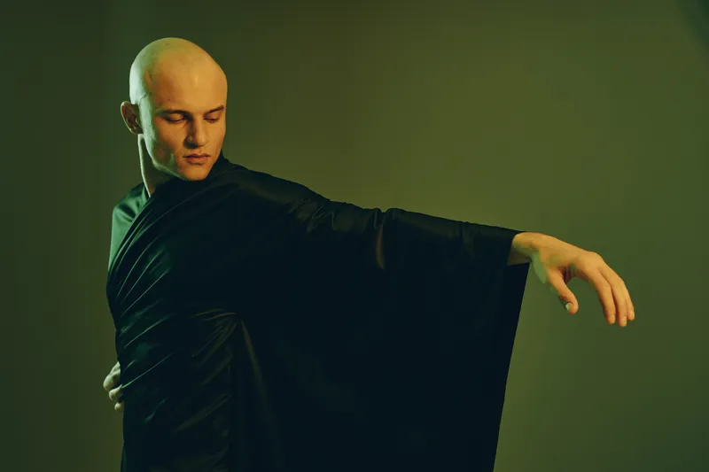 Elegant man in a flowing black robe with a serene expression, striking a graceful pose in soft, muted lighting. A captivating and unique composition highlighting fluidity.