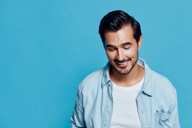 Smiling man wearing a light denim shirt looks down with a joyful expression against a blue background. Cheerful portrait capturing happiness and warmth.