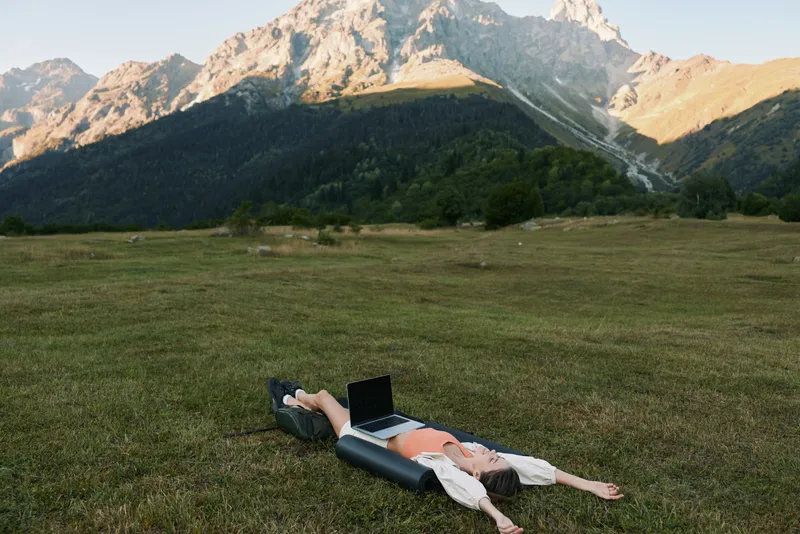 A person lies on a grassy field with a laptop beside them, enjoying a peaceful outdoor workspace surrounded by mountains as sunlight bathes the scene.