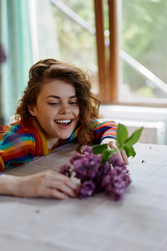 A joyful woman sits at a table indoors by a window, reaching for a bouquet of purple flowers with a bright smile, capturing a cozy home lifestyle moment full of warmth and calm