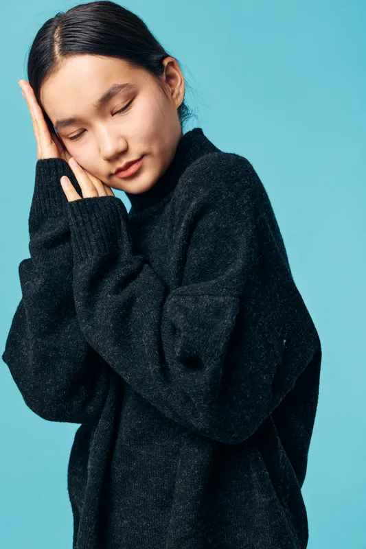 Young woman in black sweater with eyes closed standing against blue background, posing with hands gently touching her face, portrait with calm and peaceful expression.