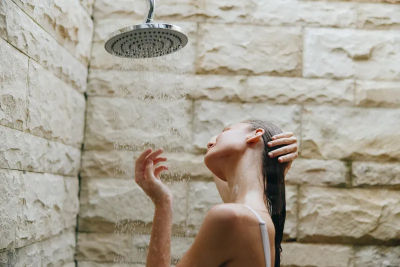 Woman showering under outdoor rain shower with natural stone wall background, wet hair, relaxed mood, and soft daylight ambiance.
