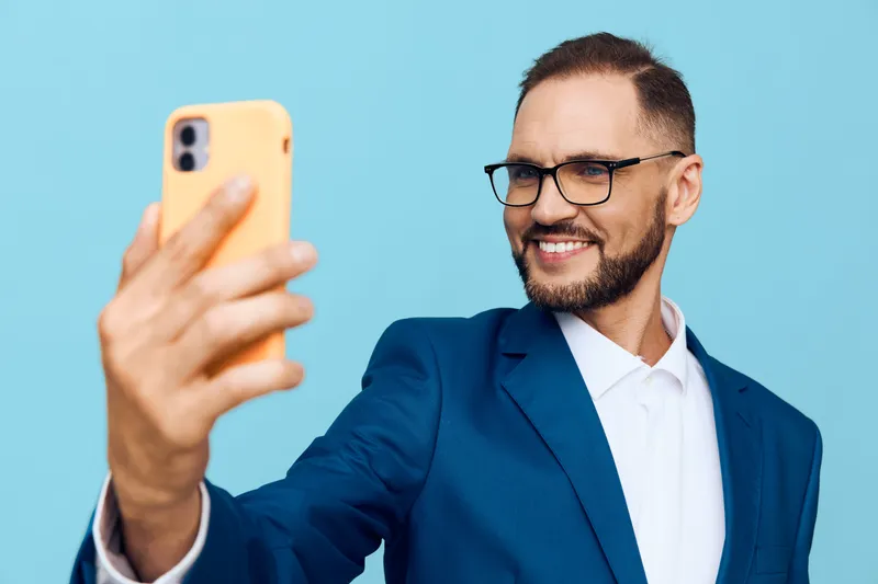 A professional man in a blue suit smiles confidently while taking a selfie against a solid blue background, presenting ambition, focus, and modern work life in a single frame.