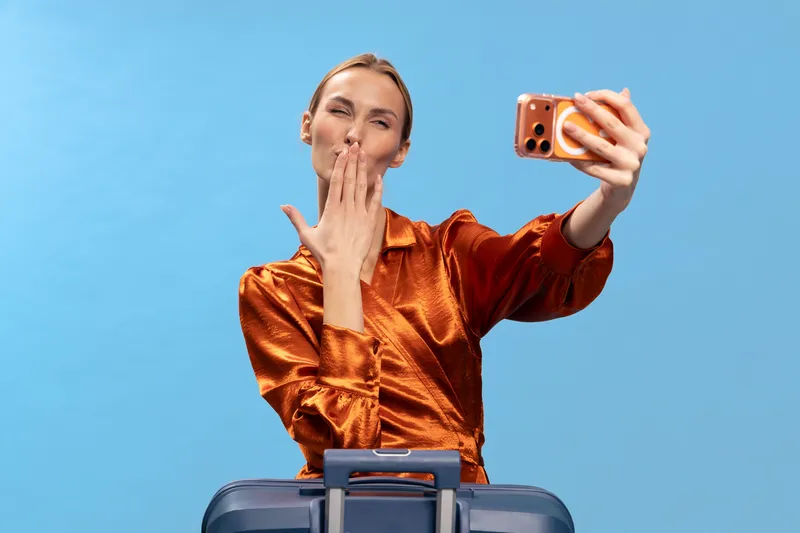 Young woman in shiny orange blouse takes selfie with smartphone while blowing kiss. Blue background and suitcase visible. Concept of travel, social media, and positive emotions.