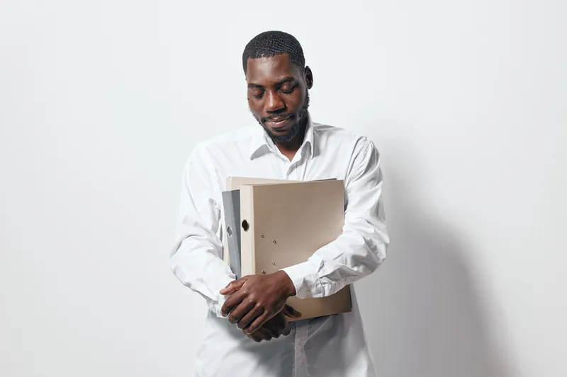 African American man with dark skin wearing white shirt holding cardboard folder and binder, standing against white background with calm expression and closed eyes.