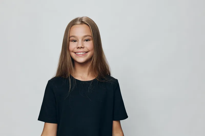Girl child with long hair wearing black t shirt smiling and looking at camera on grey background. Happy child portrait with casual clothes and natural expression.