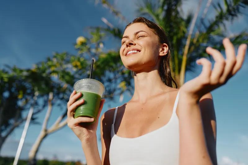 Smiling woman enjoying a green smoothie outdoors, sunlight on her face, white tank top, vibrant greenery in the background, lifestyle moment focused on health and wellness, bright.