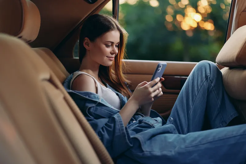 Young woman in casual denim outfit sitting in a car enjoying her phone, conveying relaxation and freedom in a tranquil setting.