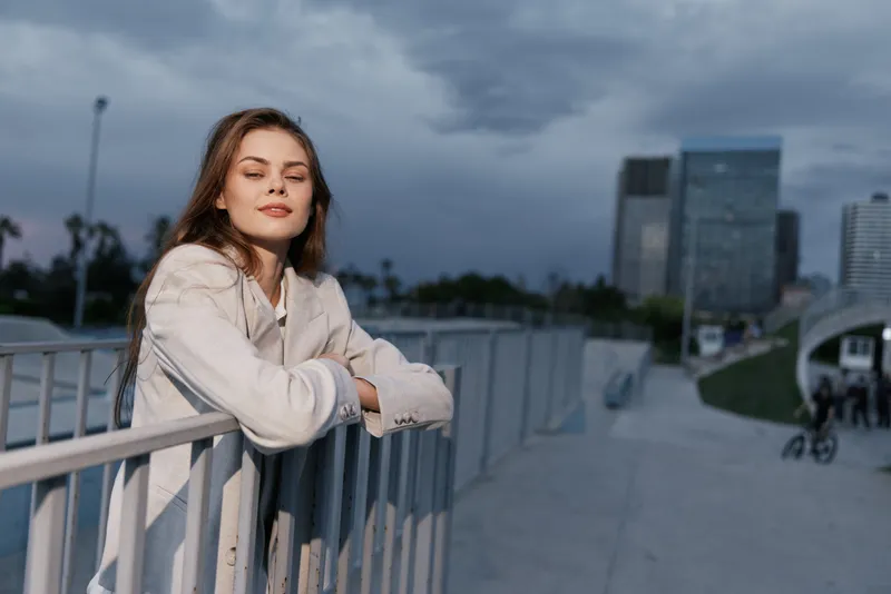 Woman in light beige coat leaning on railing with closed eyes in urban outdoor setting under moody sky, casual style, calm emotion, business concept