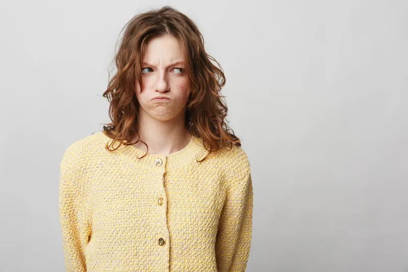 Young woman with curly hair wearing yellow sweater looks confused and displeased, isolated on grey background. Concept of doubt, frustration, and negative emotions in casual portrait.