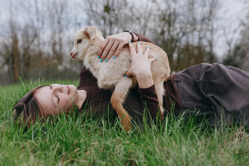 Young woman lies on green grass holding a small goat in her arms enjoying a peaceful moment outdoors in casual clothes during a lifestyle photoshoot.
