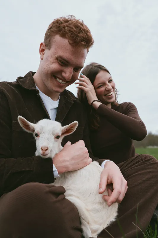Cheerful couple outdoors, holding a young goat kid, warm sunlight, relaxed pose, soft breeze, natural colors, joyful rural moment in a field