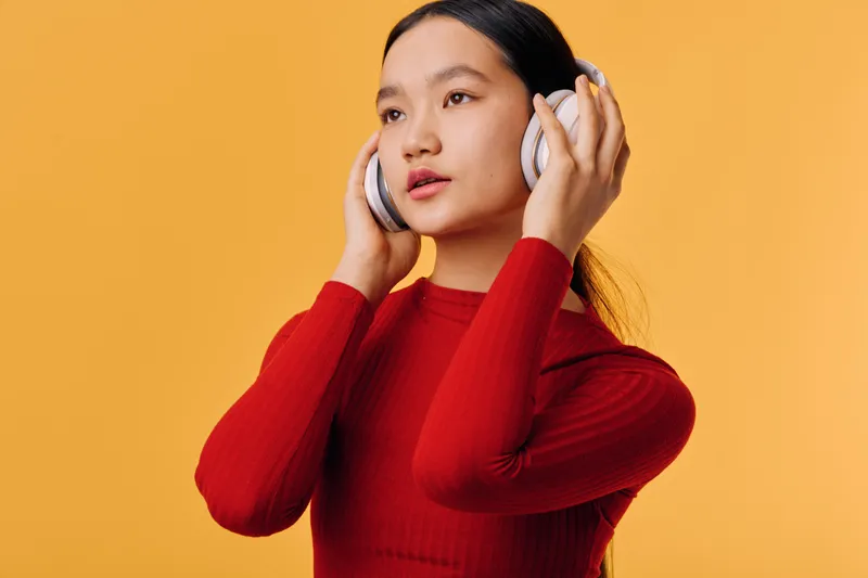 Young woman wearing red sweater listens to music with wireless headphones against yellow background. Portrait of female with long dark hair enjoying sound with eyes open and hand on earphone.