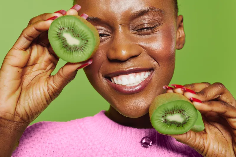 Happy African woman with dark skin holds two halves of fresh kiwi fruit near her face smiling with closed eyes on green background. Concept of healthy food, wellness, and natural beauty.