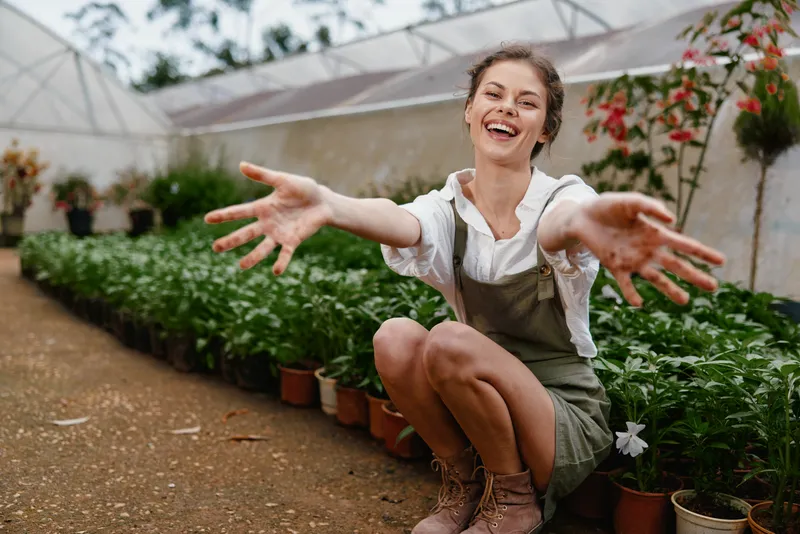 Young woman joyfully embracing nature, wearing a white shirt and overalls, surrounded by plants in a greenhouse, capturing the essence of gardening and happiness