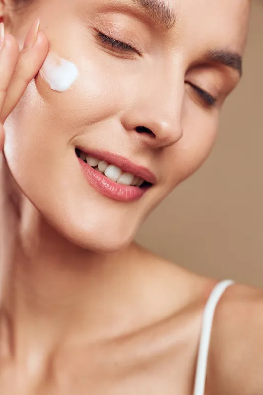 Smiling woman with clear skin applying white face cream on cheek, close-up with closed eyes on beige background. Concept of skincare, beauty, wellness, and self-care routine.