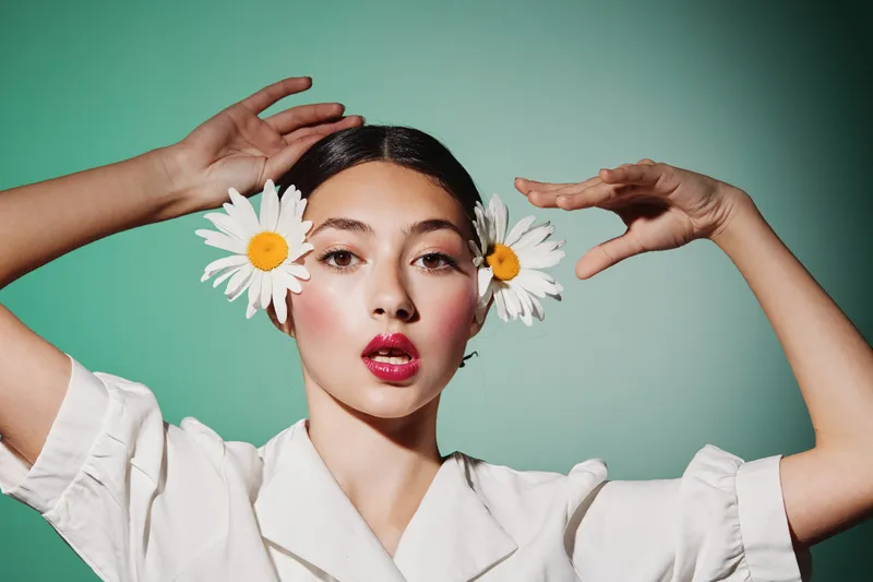 Young woman with creative makeup and two fresh daisies in her hair poses with raised arms on green background. Concept in beauty, fashion, editorial, and floral.