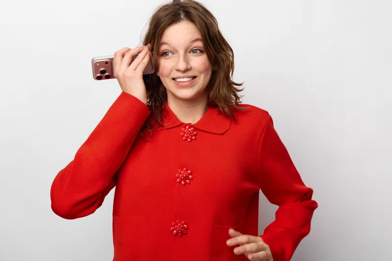 Smiling young woman in red sweater holds smartphone near ear, looking away with happy expression. Concept of communication, technology, casual lifestyle, and positive emotions.