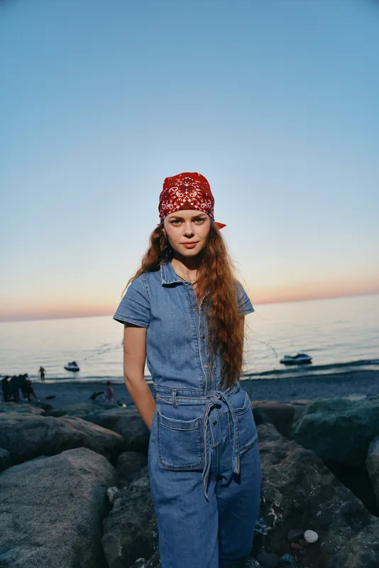 Lifestyle woman in modern boho-western denim street style wearing a red bandana by the rocky beach at sunset with warm film color tones.