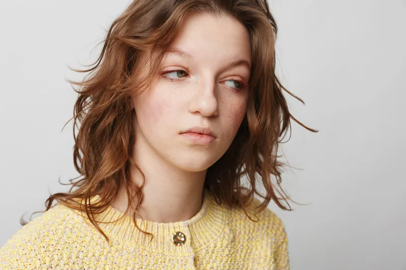 Young woman with wavy brown hair wearing yellow sweater looks away with thoughtful expression on light background. Concept of contemplation, emotion, and natural beauty.
