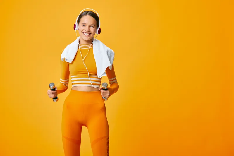 Woman in vibrant orange workout outfit with headphones, holding dumbbells and towel over shoulders in a bright studio, ready for fitness training and sport performance.