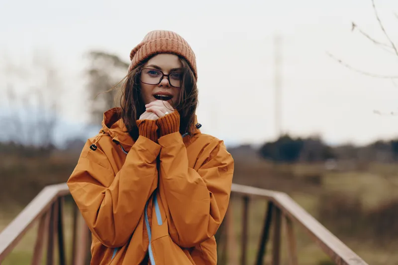 Woman lifestyle portrait outdoors in winter, wearing a yellow coat, brown beanie and glasses, she smiles and touches her chin on a bridge walkway, golden hour light and calm landscape blur behind