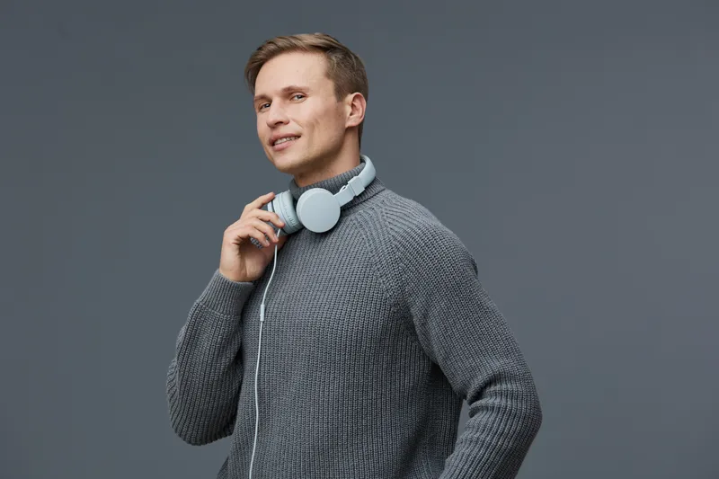 Smiling man wearing gray sweater and white headphones holding headphone cable on gray background studio portrait. People lifestyle concept