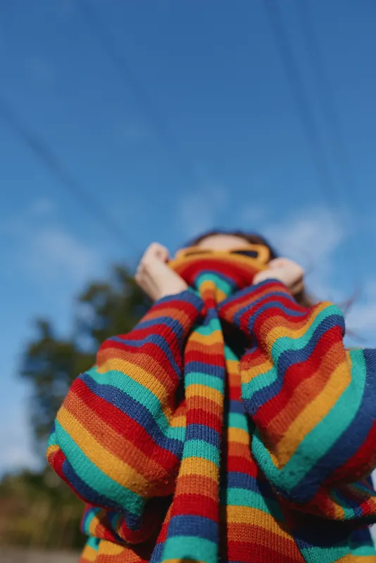 Child wearing a rainbow sweater looking up at blue sky outdoors, playful and smiling in a casual outfit. Low angle portrait with vibrant knit and sunny daylight.