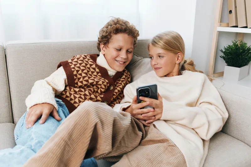Two children sit close on a soft sofa, smiling as they share a smartphone screen, wrapped in cozy sweaters and a warm blanket, enjoying a cheerful, relaxed home moment.