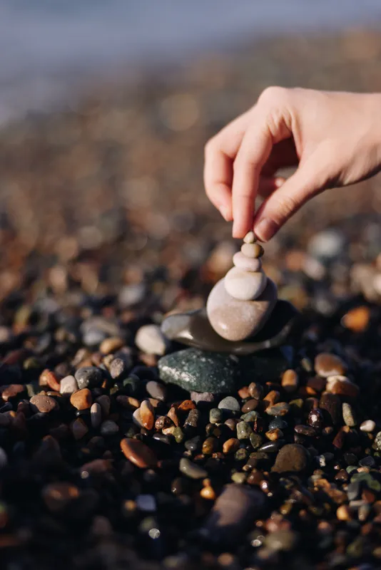 Stones balance on a pebble beach as a hand carefully stacks a small cairn, creating a zen moment of calm, focus, and natural sculpture amid smooth rocks and coastal sand.