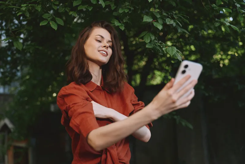 Cheerful woman outdoors, taking a selfie with a smile, warm natural light, casual red shirt, lush greenery in the background, vibrant lifestyle moment