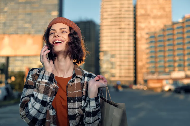 Woman shopping and talking on phone with smile in urban street during golden hour, candid authenticity and mindful living vibe, emotional storytelling of joyful city life