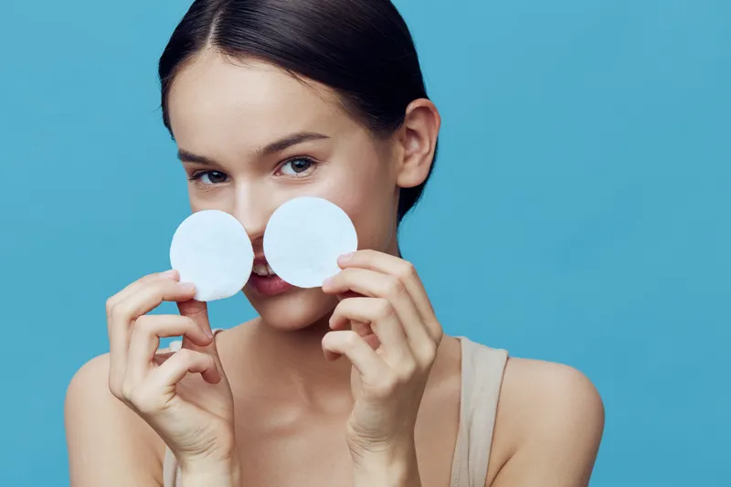 Young woman holding cotton pads in front of her face, smiling, on a blue background, representing skincare and beauty concepts