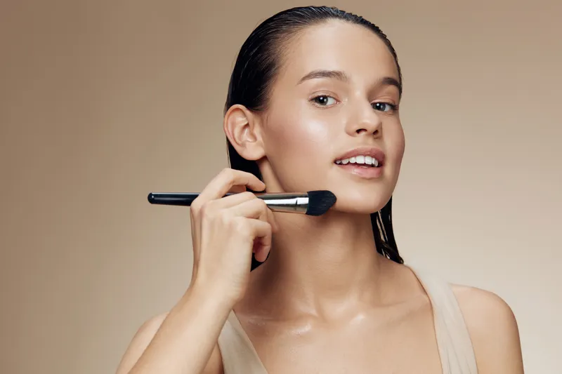 Young woman applying makeup with brush, showcasing natural beauty against a soft beige background, emphasizing healthy skin and a fresh look
